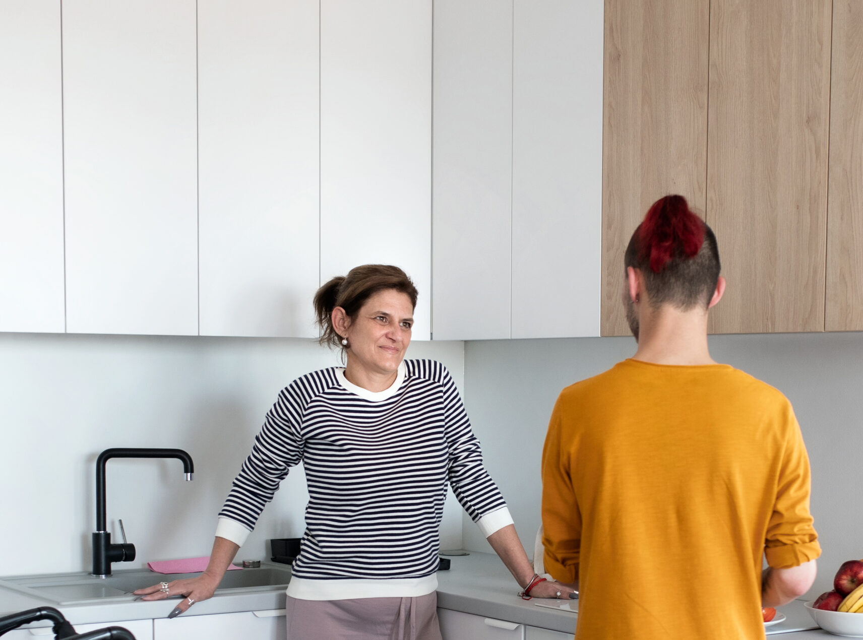 A disabled mature woman in wheelchair talking to son in kitchen indoors at home.
