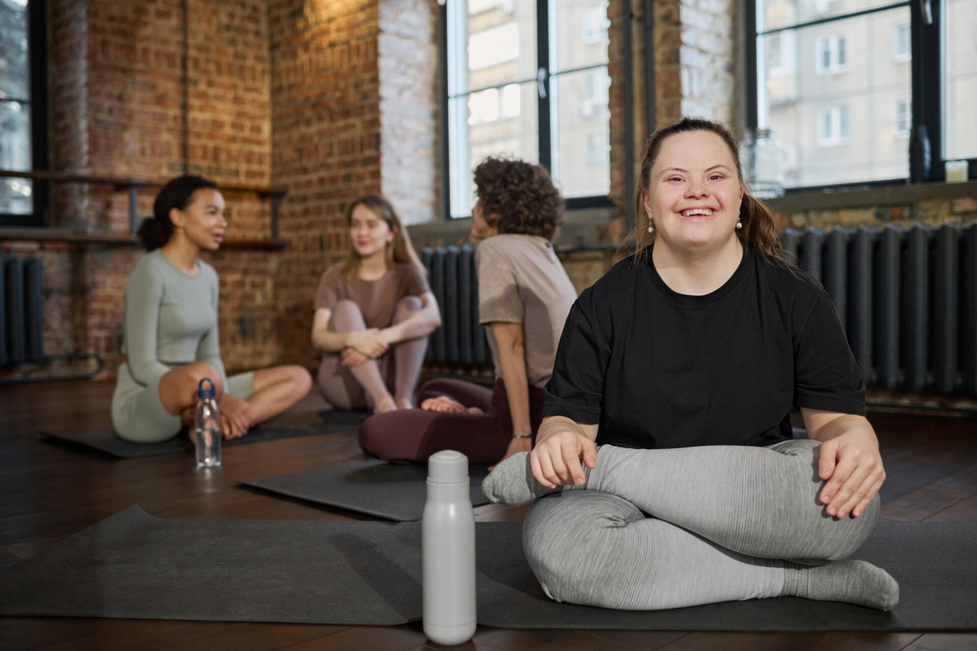 Young cheerful woman with Down syndrome in activewear sitting on mat and looking at camera against group of active girls in gym