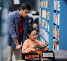 Portrait of Asian girl with disability in library choosing books with friend assisting, inclusivity concept