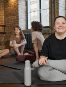 Young cheerful woman with Down syndrome in activewear sitting on mat and looking at camera against group of active girls in gym