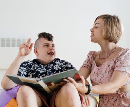 Children with disability reading book and communicating. Cerebral palsy boy talking with woman tale therapist. Rehabilitation center, mother with teenage person learning to speak, pronounce at home