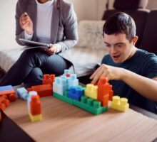 Happy down syndrome man playing with toy blocks and having fun with psychologist during home visit.